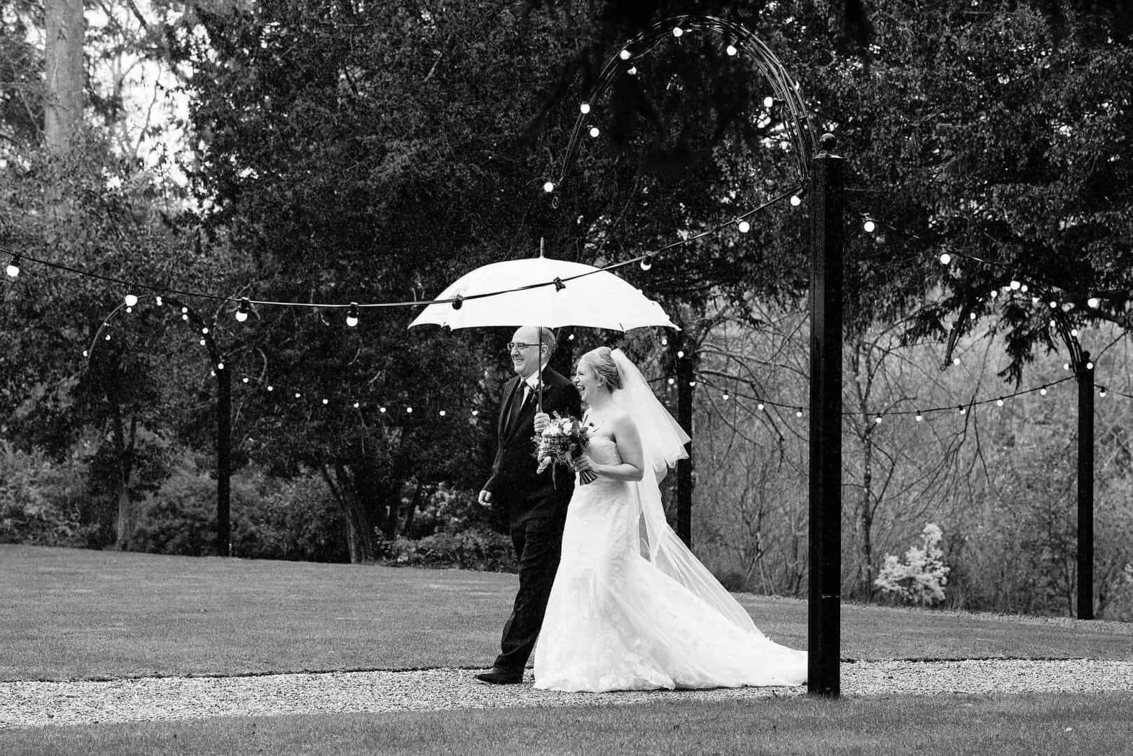 Bride arrives in the rain with her dad under a big umbrella at Larmer tree gardens